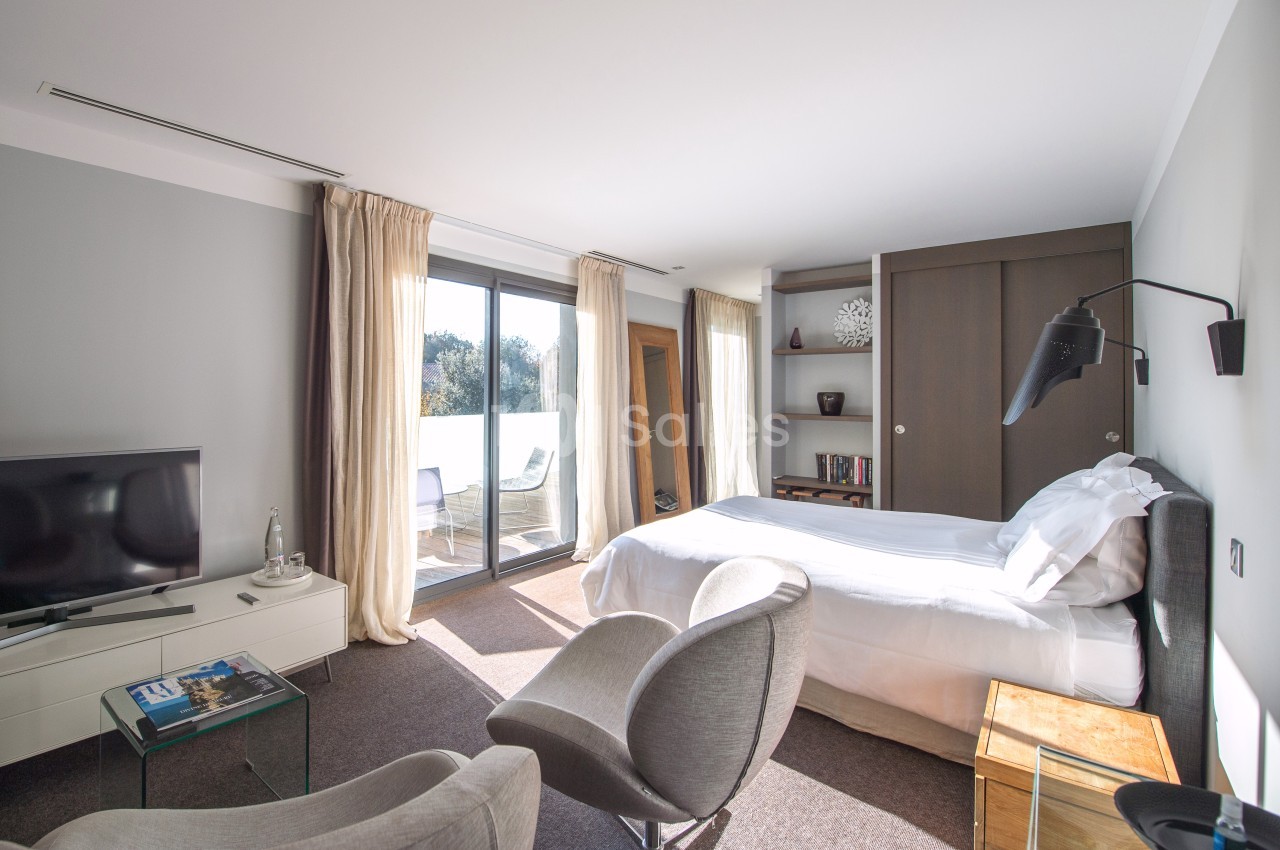 Chambre lumineuse avec lit double, fauteuils, télévision, grande baie vitrée et accès à un balcon.
