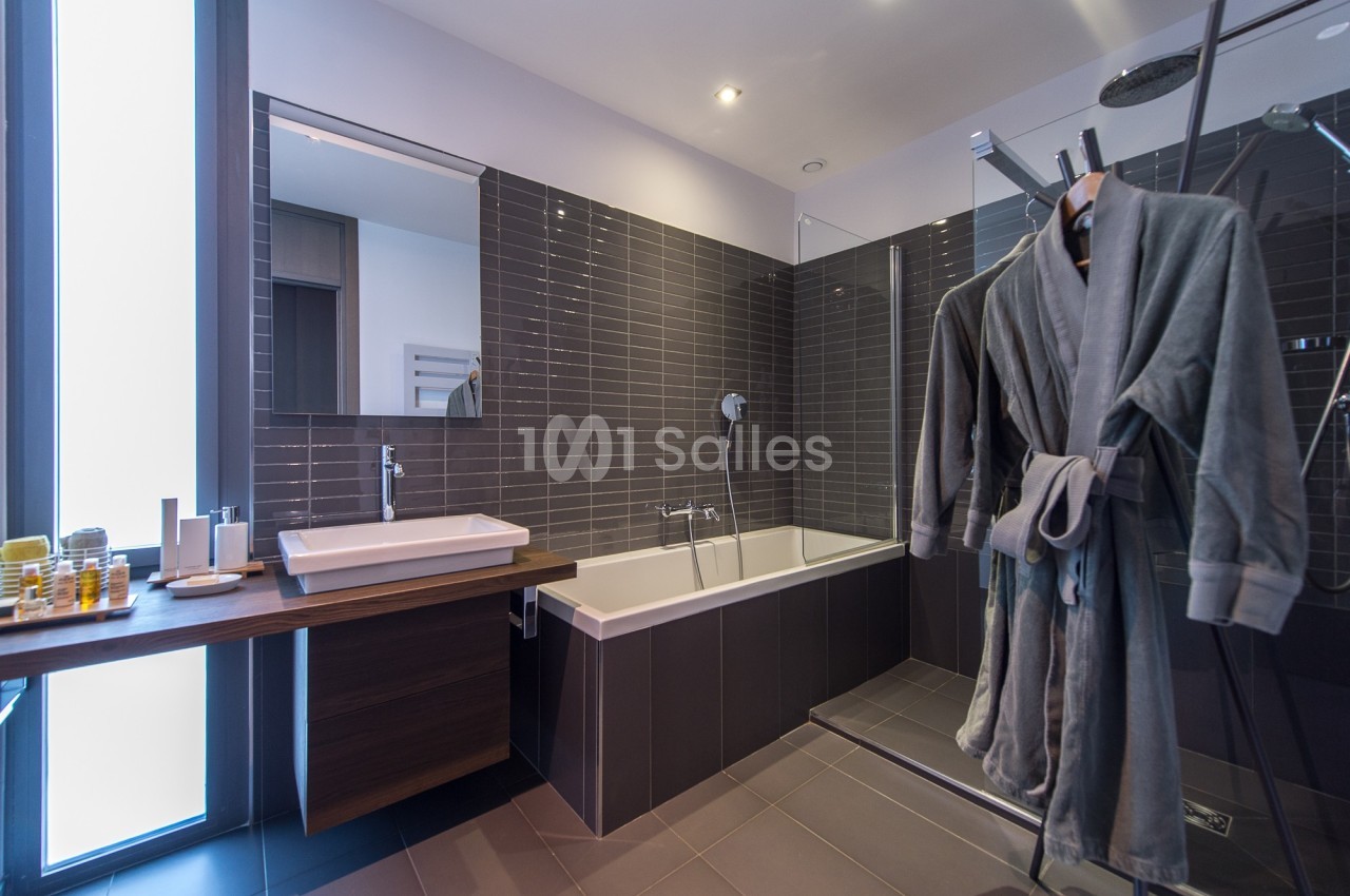 Salle de bain moderne avec baignoire, douche vitrée, lavabo et peignoirs suspendus près d'une fenêtre.