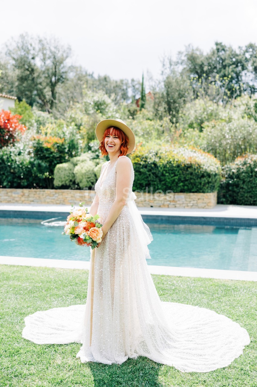 Femme souriante en robe de mariée blanche tenant un bouquet coloré, debout près d'une piscine dans un jardin.