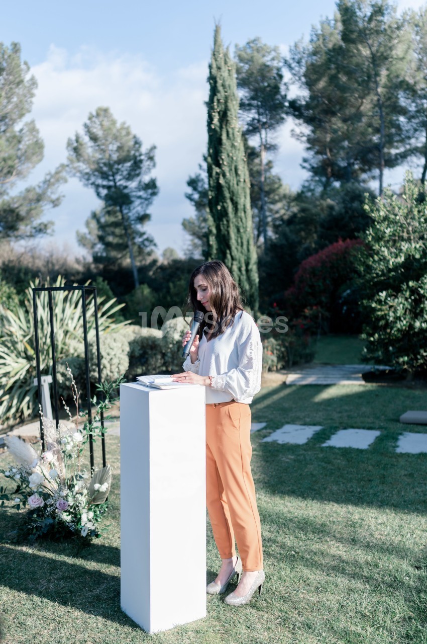 Une femme tenant un micro parle debout derrière un pupitre blanc dans un jardin arboré.