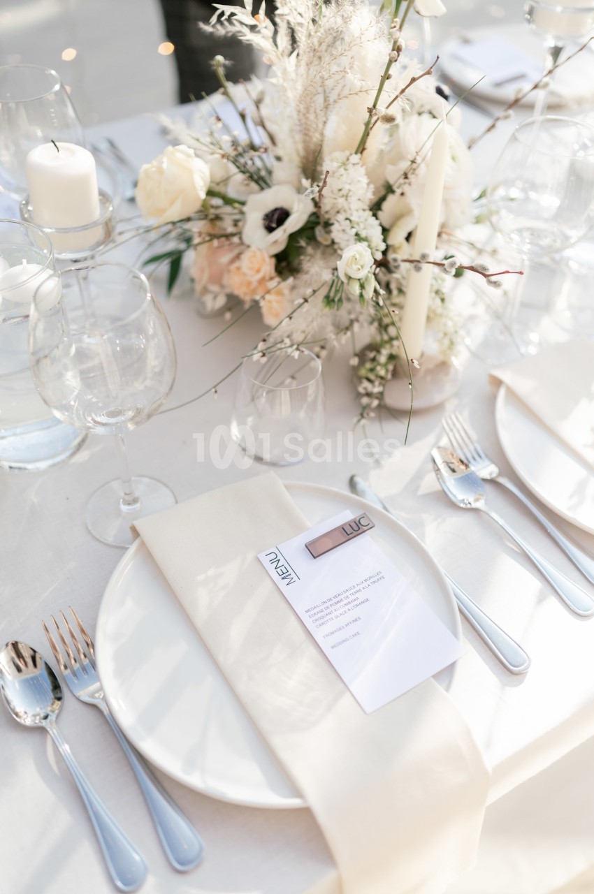 Table dressée avec élégance, comprenant des assiettes blanches, des couverts argentés, un menu et un centre de table floral.