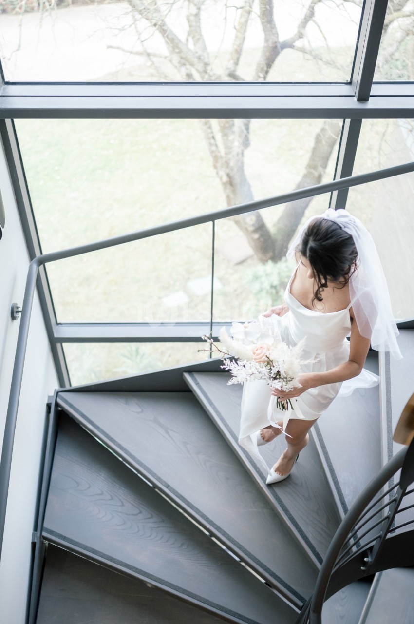 Une mariée en robe blanche et voile monte un escalier en colimaçon, tenant un bouquet de fleurs pastel.