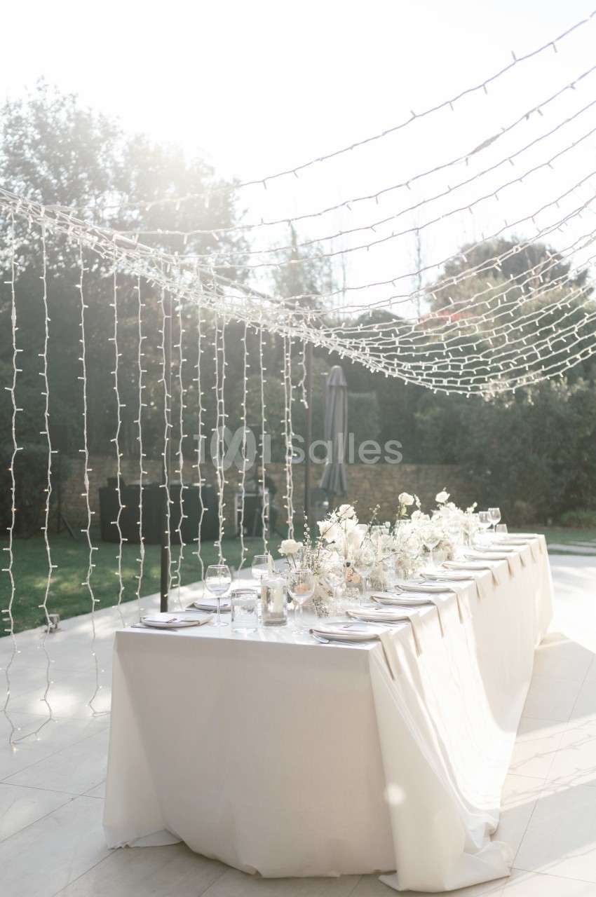 Table élégamment dressée en extérieur, décorée de fleurs blanches et éclairée par des guirlandes lumineuses suspendues.