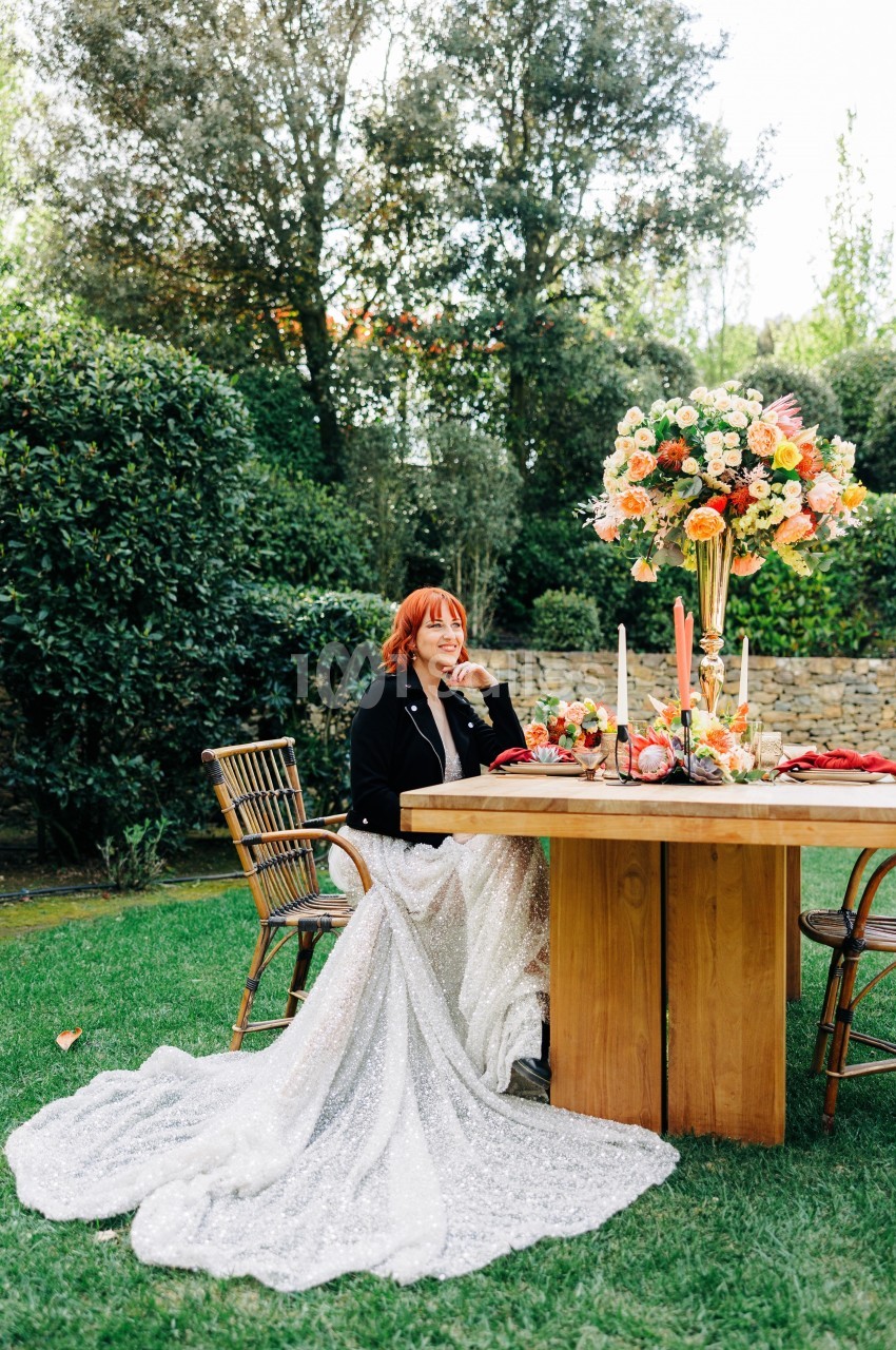 Une femme en robe blanche assise à une table décorée de fleurs colorées dans un jardin verdoyant.