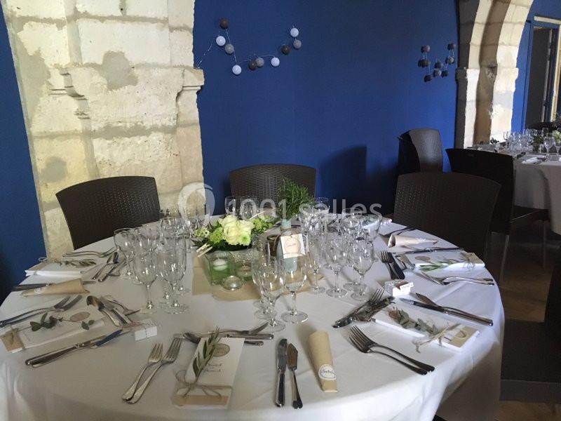 Table ronde élégamment dressée avec nappes blanches, couverts, verres et décoration florale dans une salle aux murs bleus.