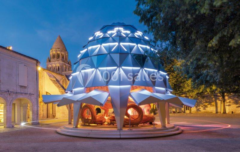 Structure géométrique illuminée en forme de dôme, installée sur une place, avec une église en arrière-plan.