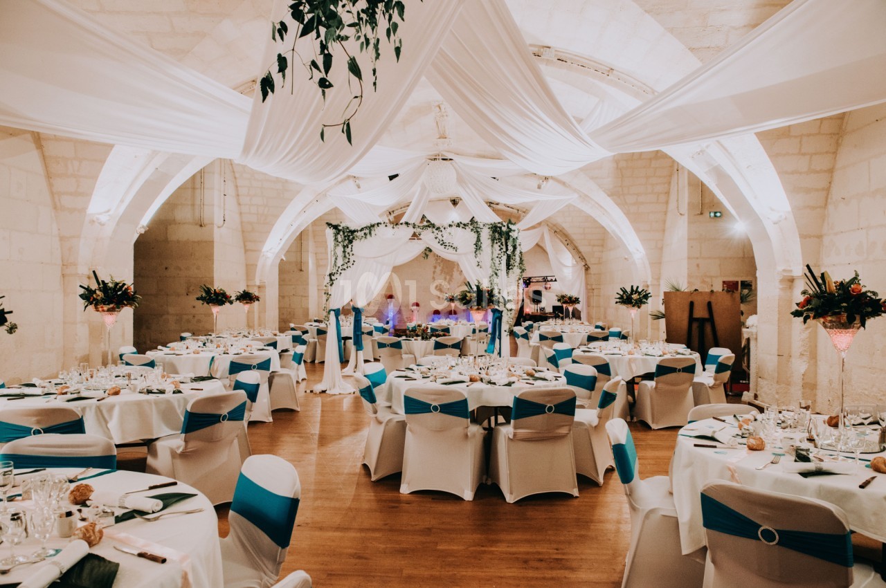 Salle de réception décorée avec des tables rondes, nappes blanches, chaises ornées de rubans bleus et plafond drapé.