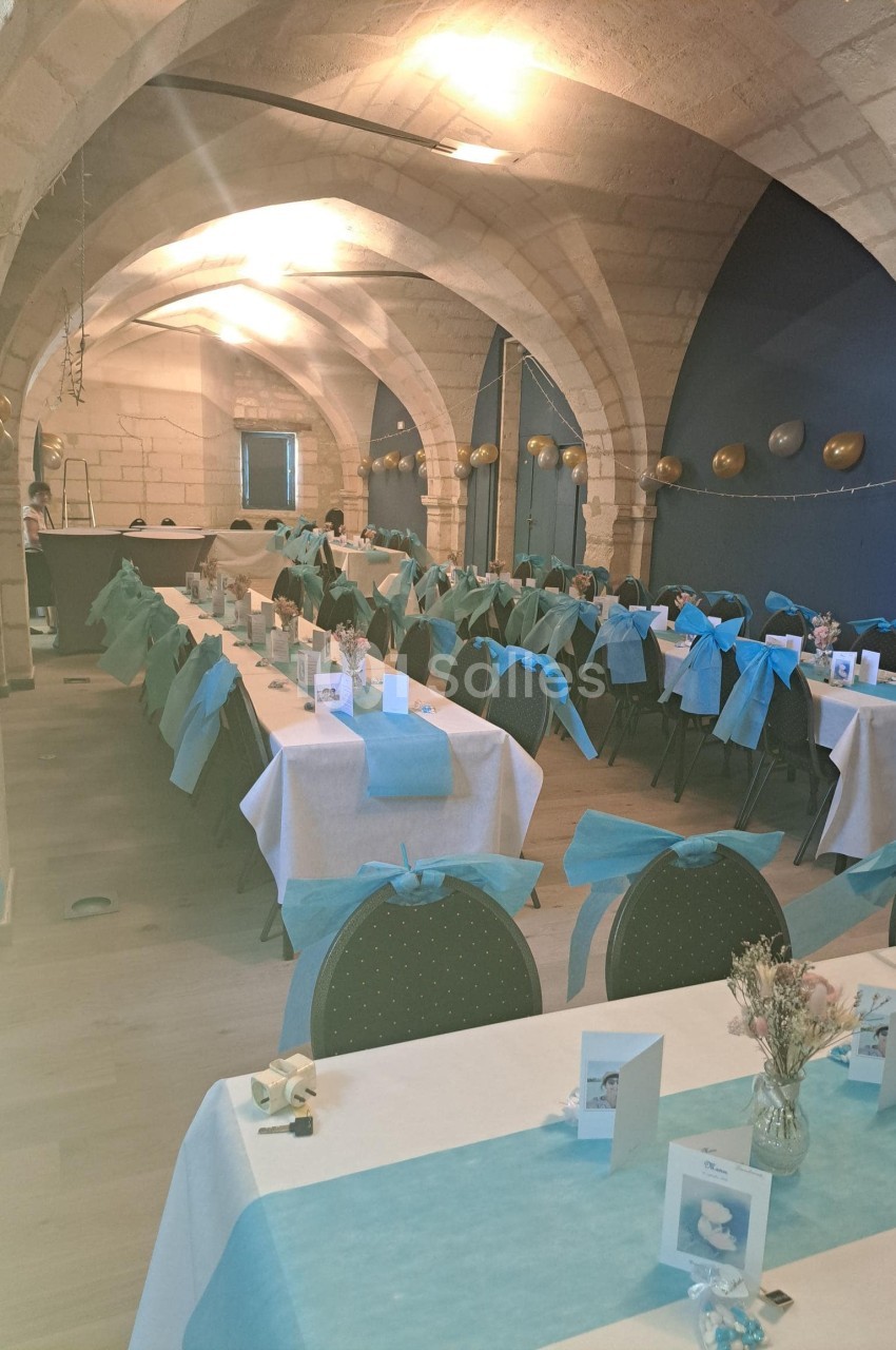 Salle voûtée décorée pour un événement, avec des tables alignées, nappes blanches et bleues, et chaises assorties.