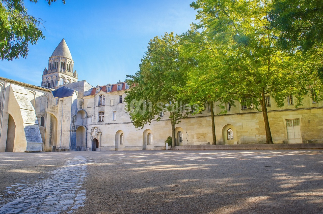 Cour pavée bordée d'arbres et bâtiments historiques, avec un clocher en arrière-plan sous un ciel bleu.