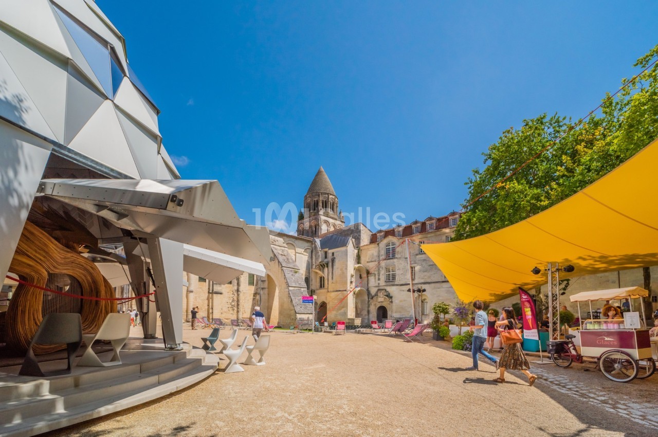 Place ensoleillée avec structures modernes, église historique en arrière-plan et stands colorés.