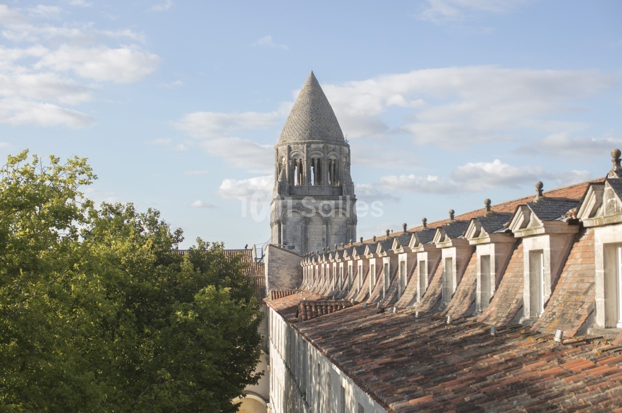 Clocher en pierre d'une église romane entouré de toits en tuiles et d'arbres sous un ciel partiellement nuageux.