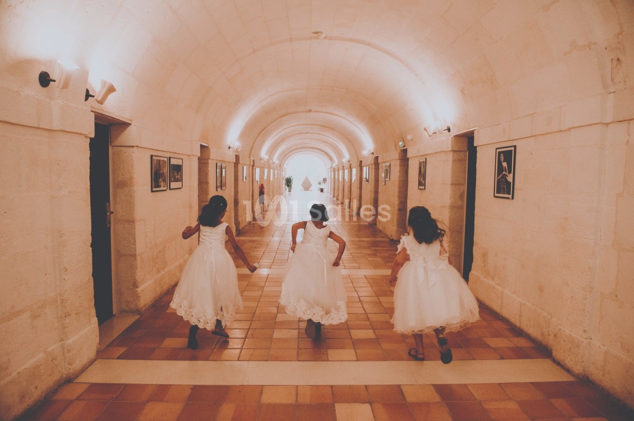 Trois jeunes filles en robes blanches courent dans un long couloir voûté éclairé par des appliques murales.