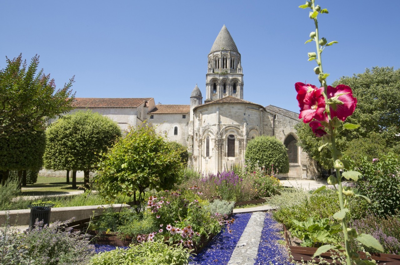 Jardin fleuri avec allée colorée devant une église romane à clocher, sous un ciel bleu dégagé.