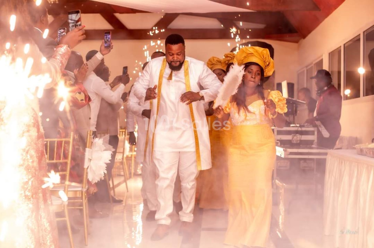 Un couple en tenue traditionnelle marche au milieu d'étincelles lors d'une célébration festive en intérieur.
