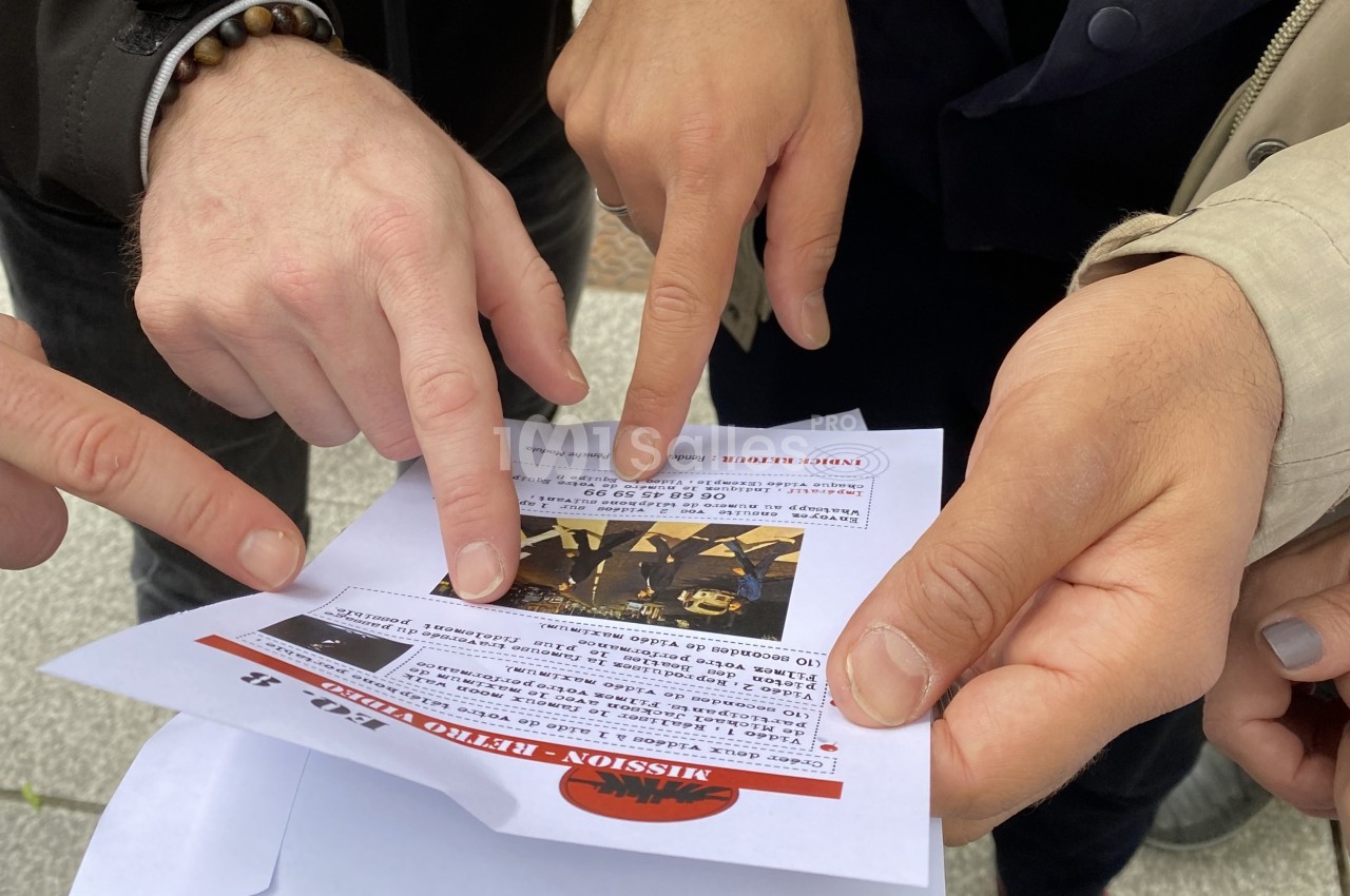 Des mains pointent du doigt des informations sur une feuille imprimée, en extérieur sur un sol pavé.