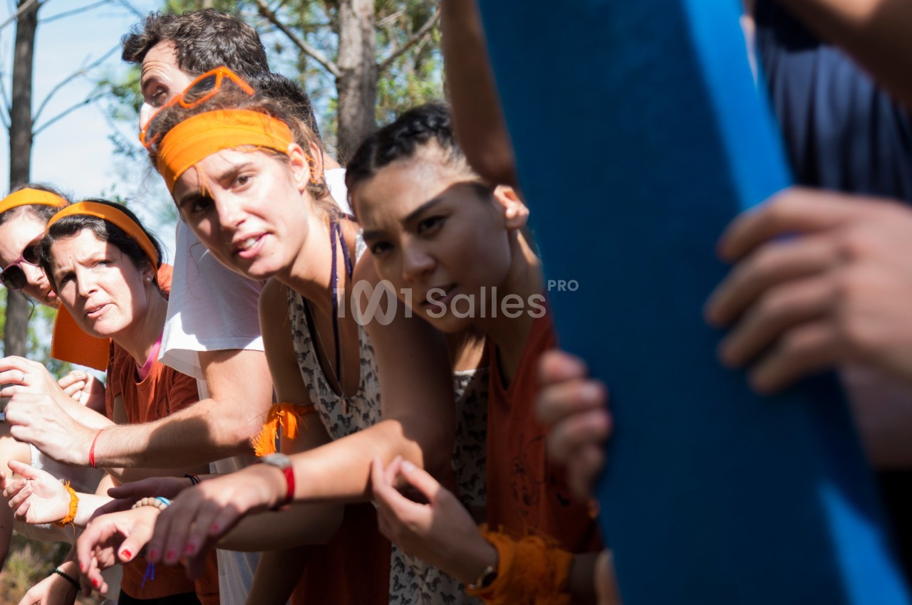 Des personnes en groupe, portant des bandeaux orange, semblent concentrées lors d'une activité en plein air.