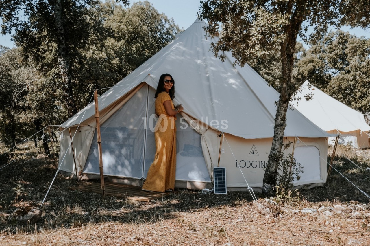 Femme en robe jaune devant une tente blanche installée dans un cadre naturel boisé.