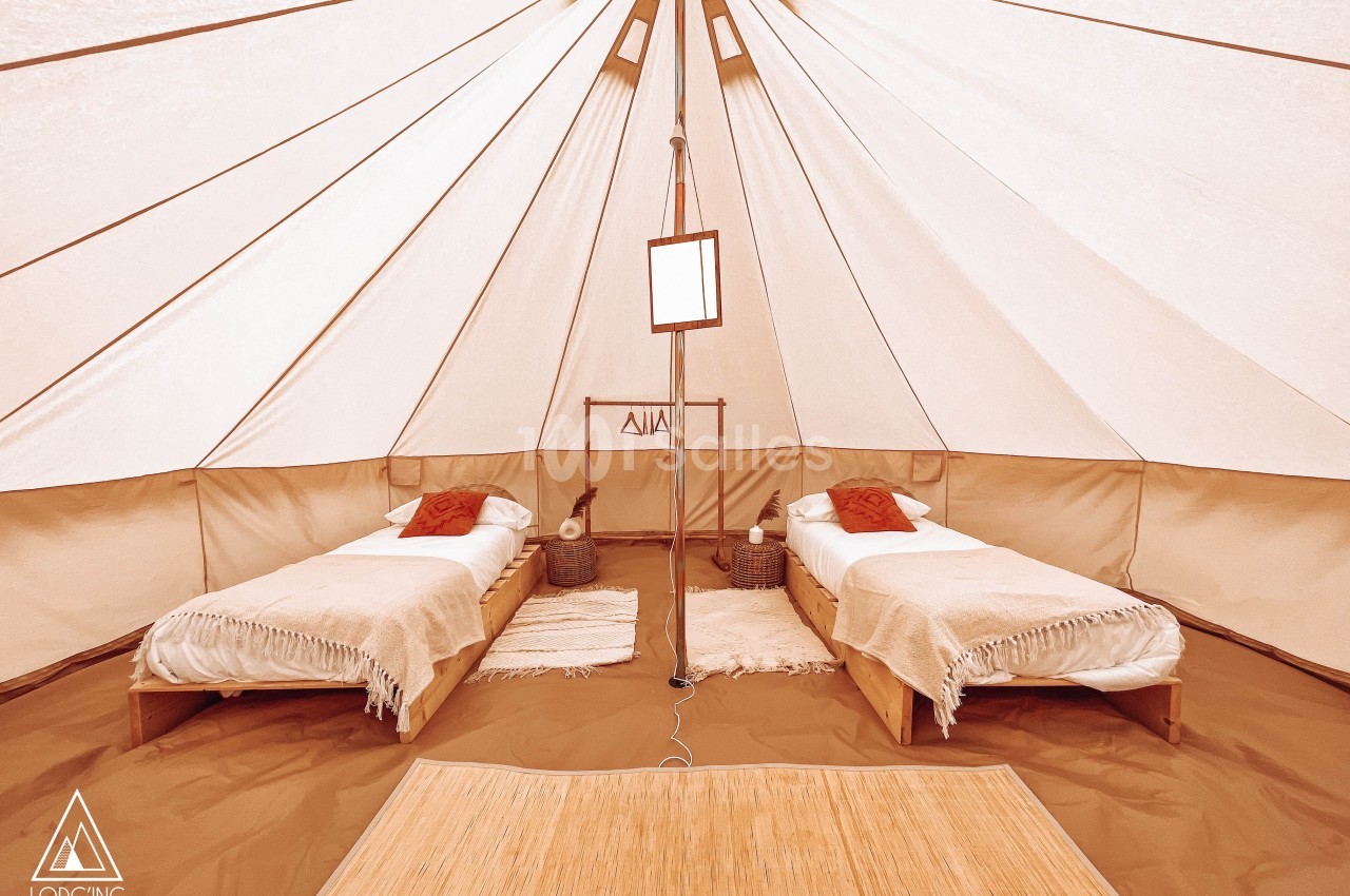 Intérieur d'une tente spacieuse avec deux lits simples, linge beige, coussins rouges et sol recouvert de tapis.