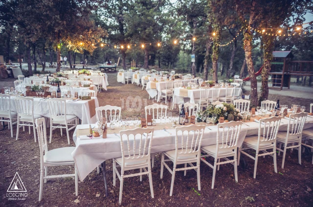 Tables dressées avec nappes blanches et chaises alignées en extérieur, sous des guirlandes lumineuses dans un cadre boisé.