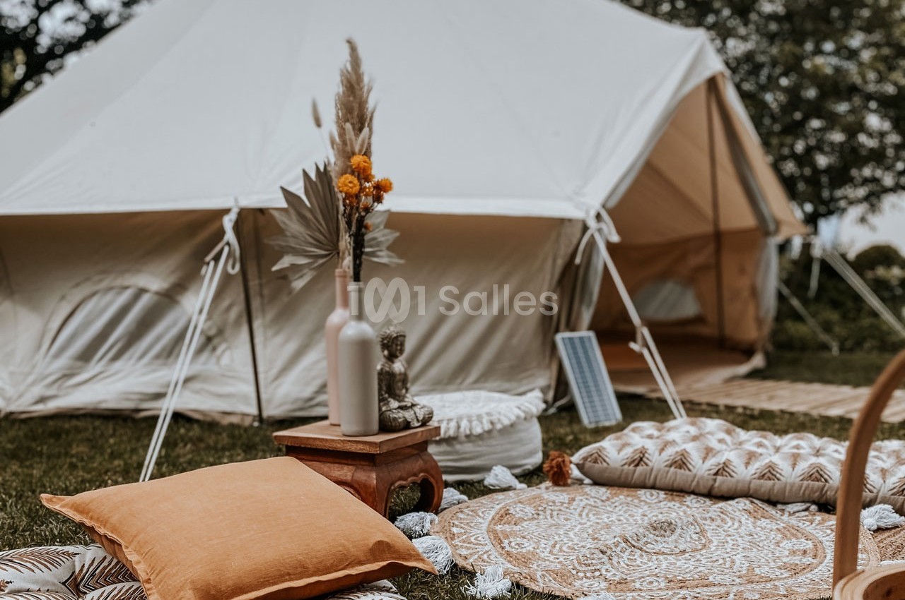 Tente beige avec décoration bohème comprenant coussins, tapis et vase sur un espace herbeux en extérieur.