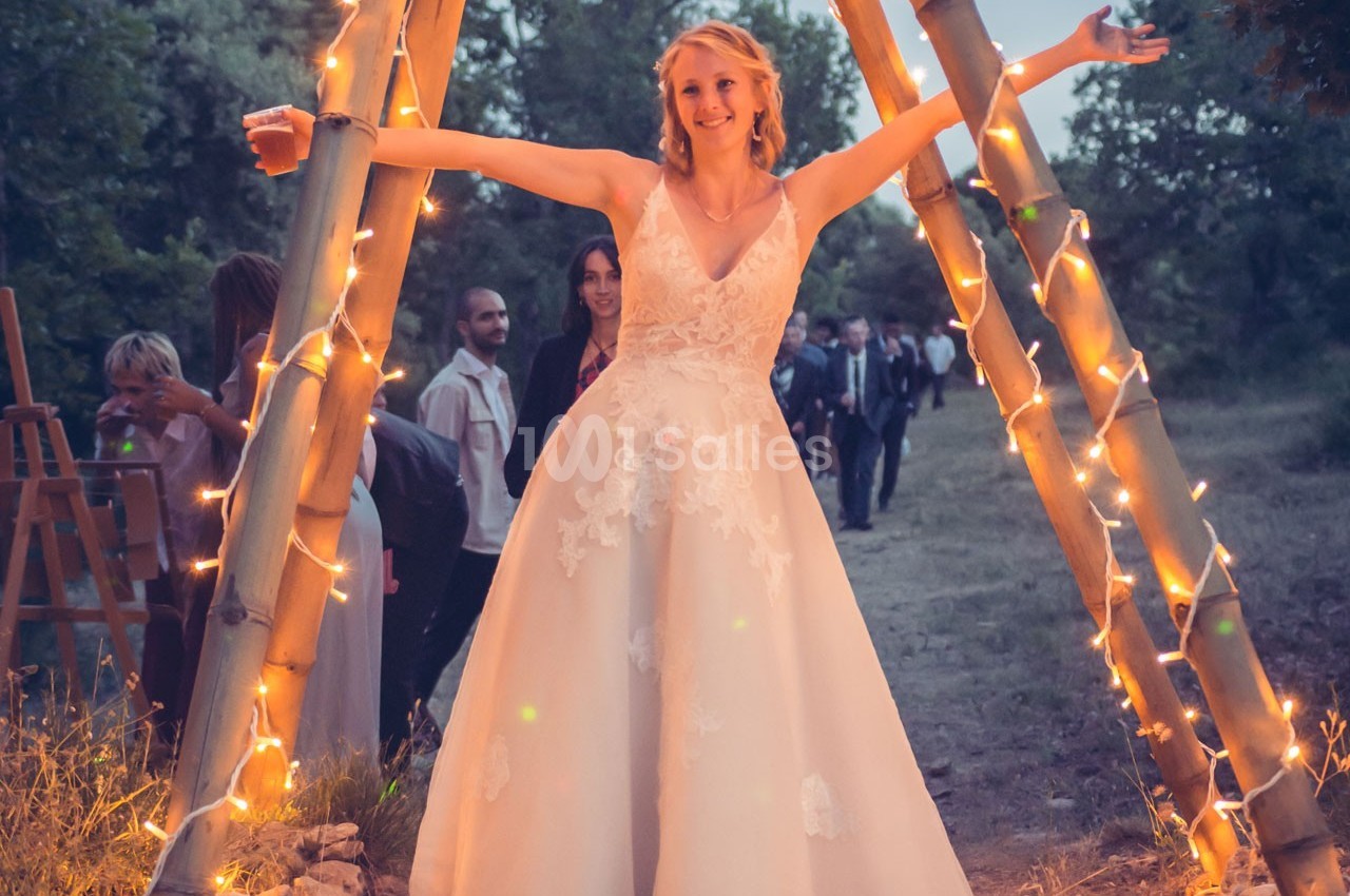 Une femme en robe de mariée sourit, bras ouverts, sous une arche décorée de lumières dans un cadre extérieur boisé.
