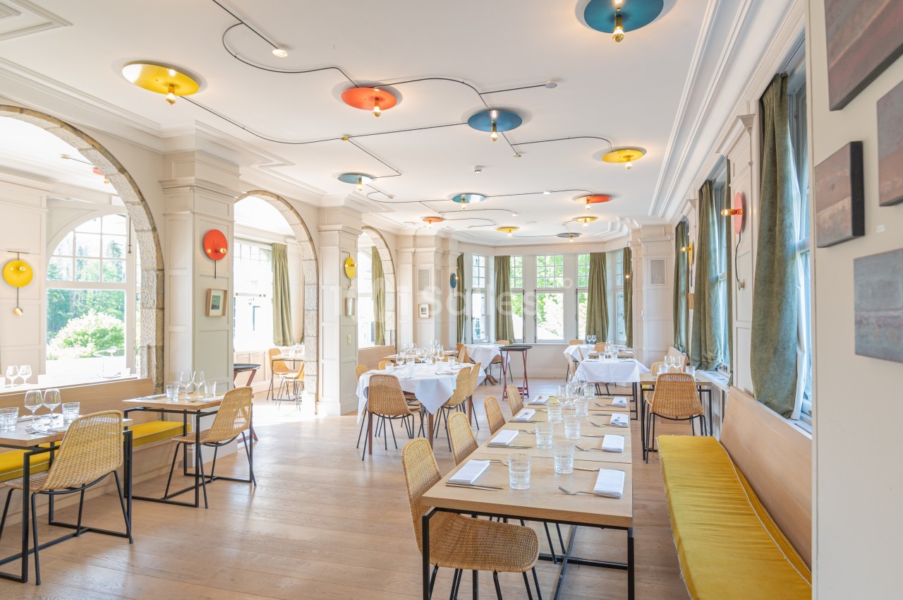 Salle de restaurant lumineuse avec grandes fenêtres, tables dressées, chaises en rotin et banquettes jaunes.