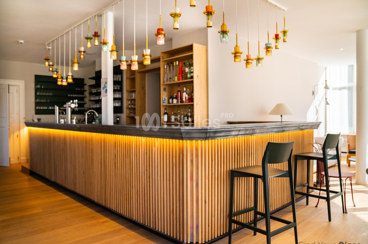 Bar moderne avec comptoir en bois, éclairage suspendu coloré et tabourets noirs sur un sol en parquet clair.