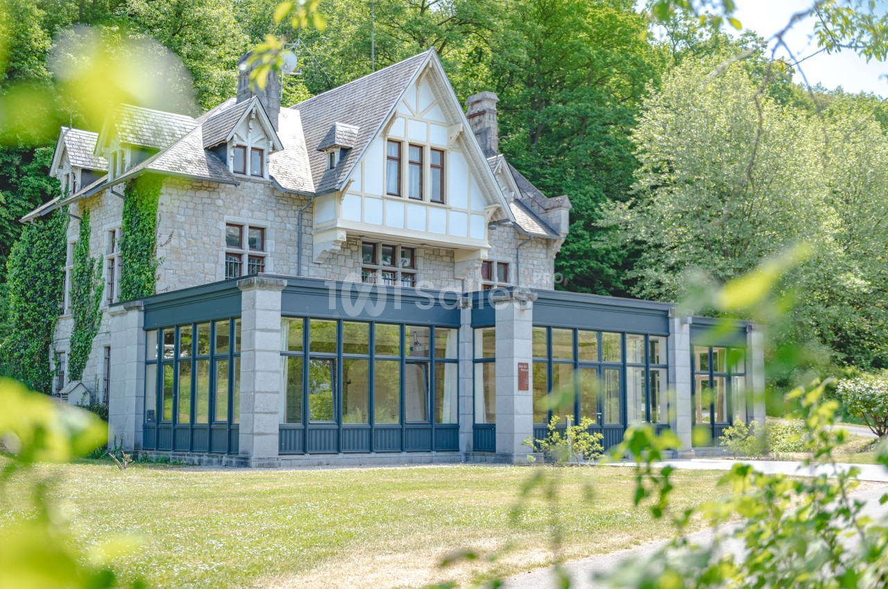 Grande maison en pierre avec véranda moderne, entourée de verdure et située dans un cadre boisé.