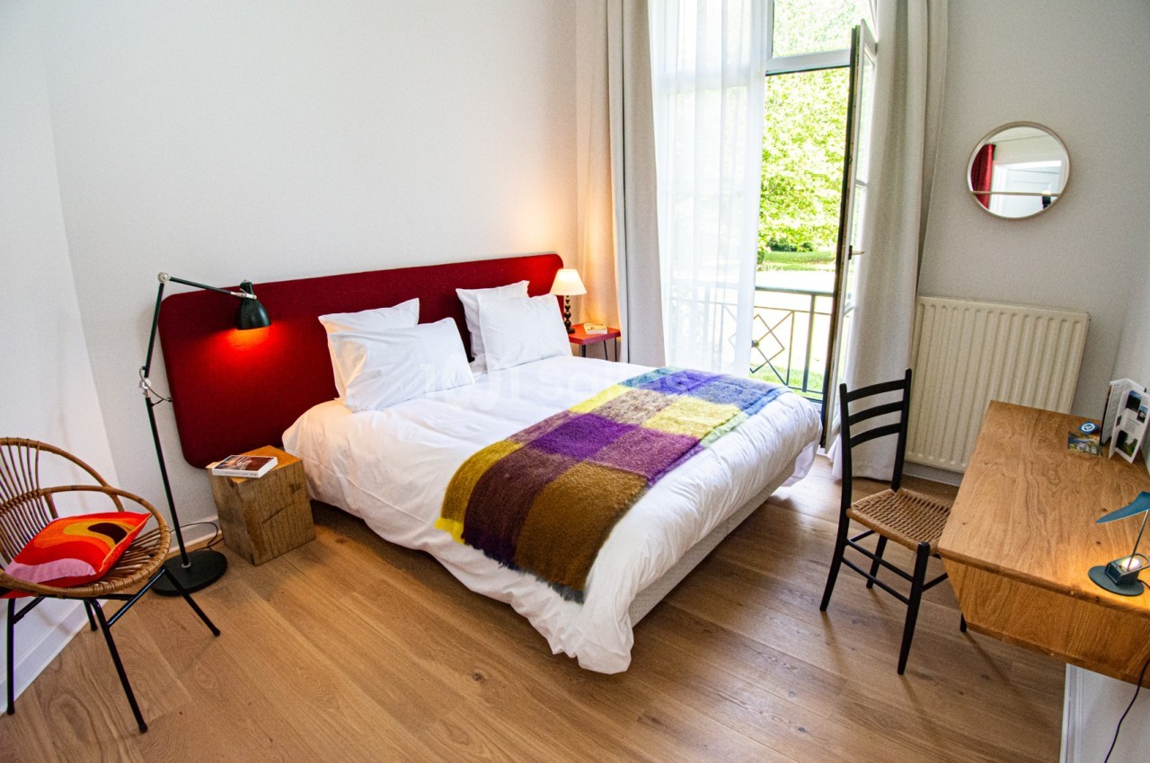 Chambre lumineuse avec lit double, plaid coloré, bureau en bois, chaise et vue sur un jardin par une fenêtre ouverte.
