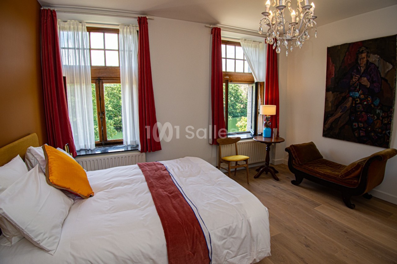 Chambre lumineuse avec lit double, fauteuil, table avec lampe, grandes fenêtres et vue sur un jardin verdoyant.