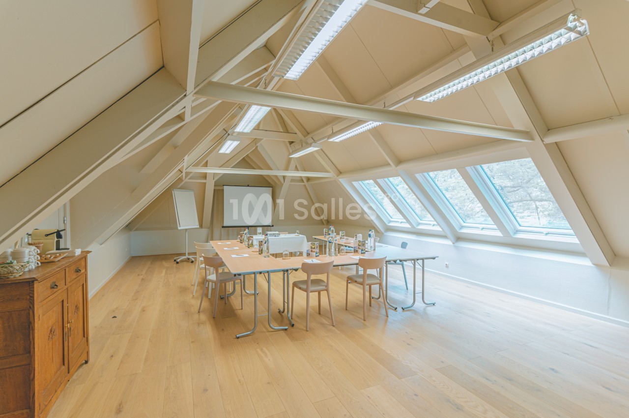 Salle de réunion lumineuse sous combles avec grandes fenêtres, table équipée de chaises et matériel de présentation.
