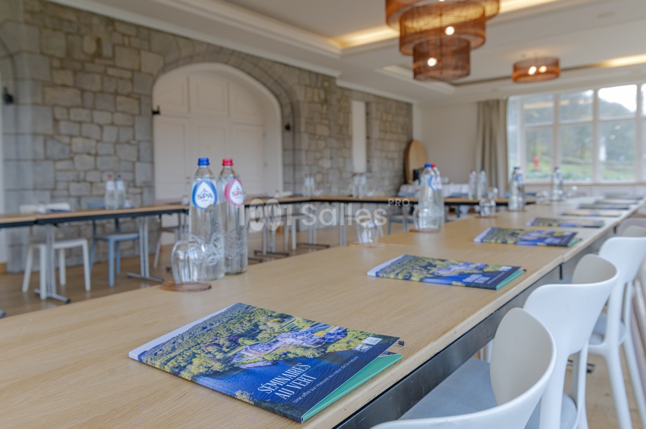 Salle de réunion lumineuse avec tables alignées, chaises blanches, bouteilles d'eau et brochures posées sur les tables.