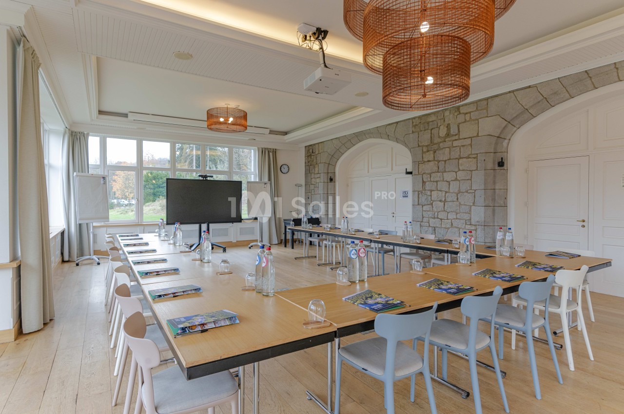 Salle de réunion lumineuse avec grandes fenêtres, tables en U, chaises colorées et écran de présentation.