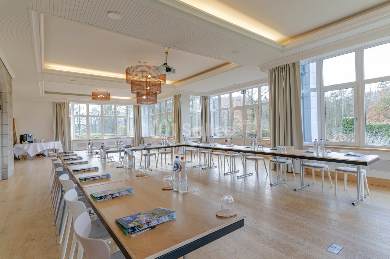 Salle lumineuse aménagée pour une réunion, avec des tables en U, des chaises, des bouteilles d'eau et des documents.