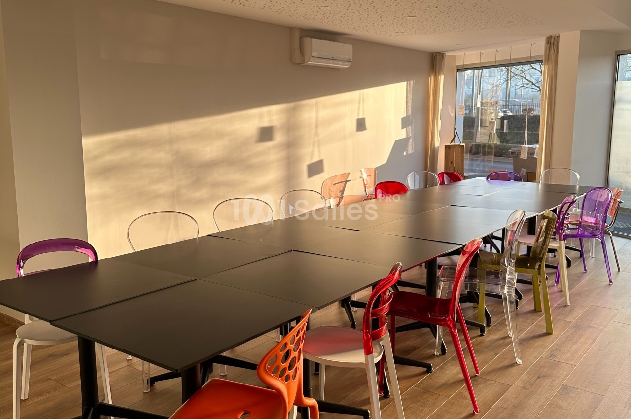 Salle de réunion lumineuse avec une grande table noire entourée de chaises colorées, près d'une fenêtre.