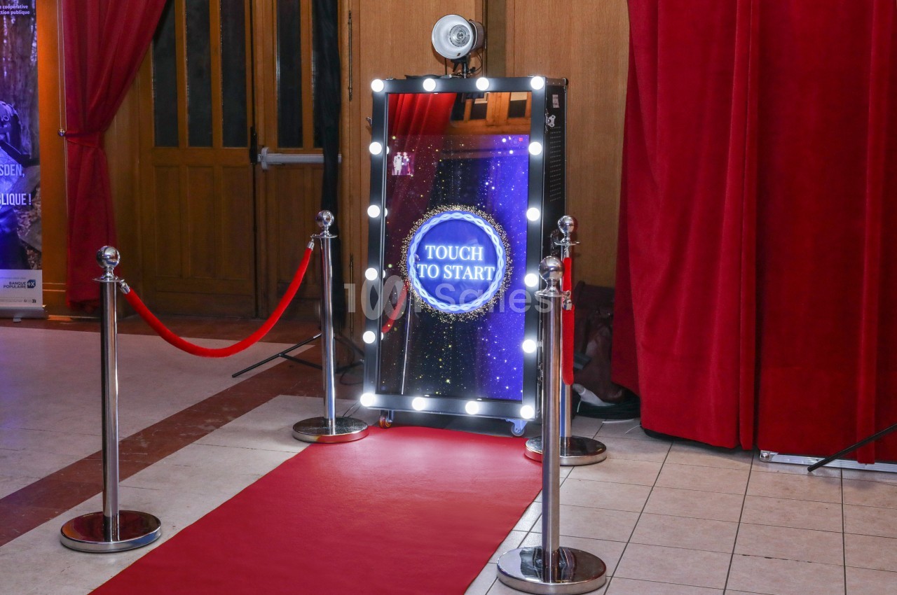 Miroir interactif lumineux avec écran tactile, entouré de rideaux rouges et d'un tapis rouge dans un espace intérieur.