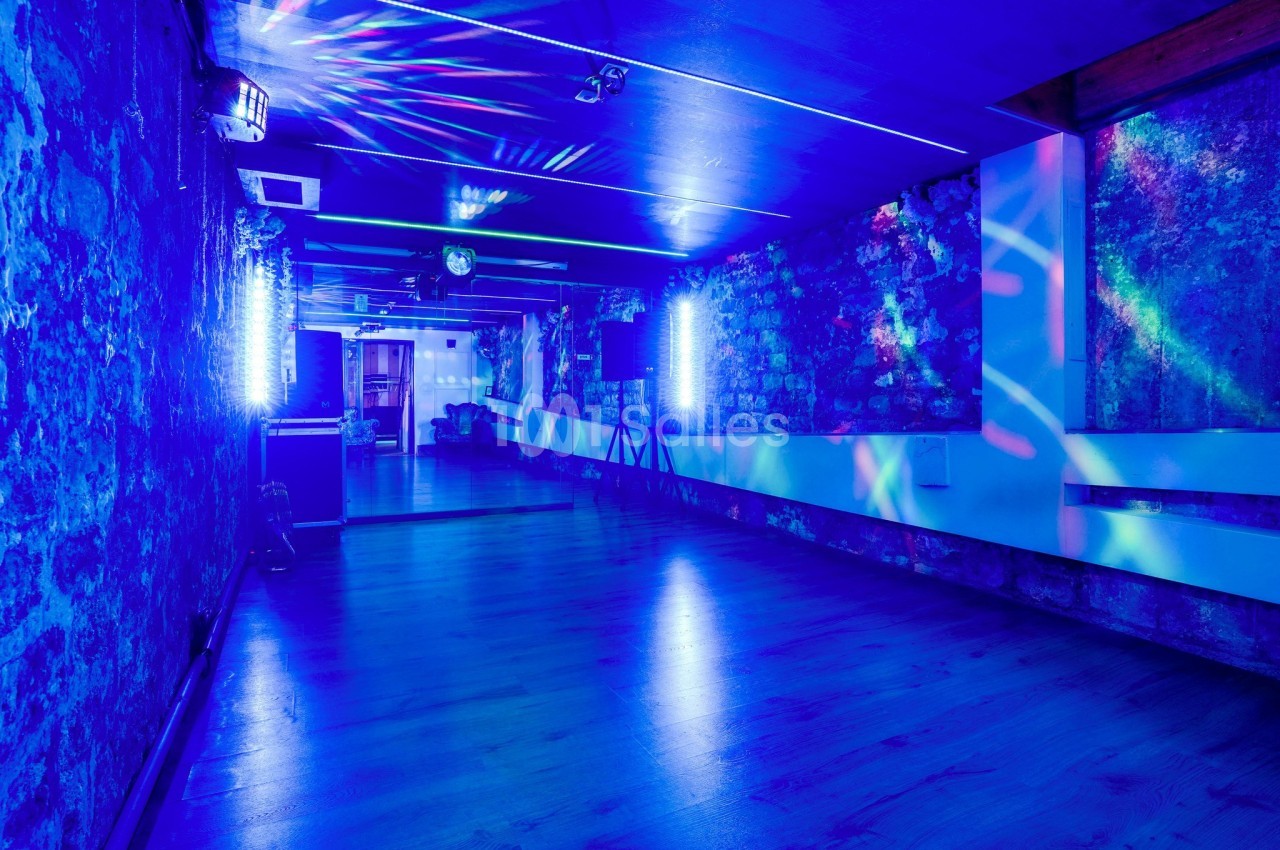 Salle avec murs en pierre, éclairage bleu et jeux de lumière colorés, sol en bois et ambiance festive.