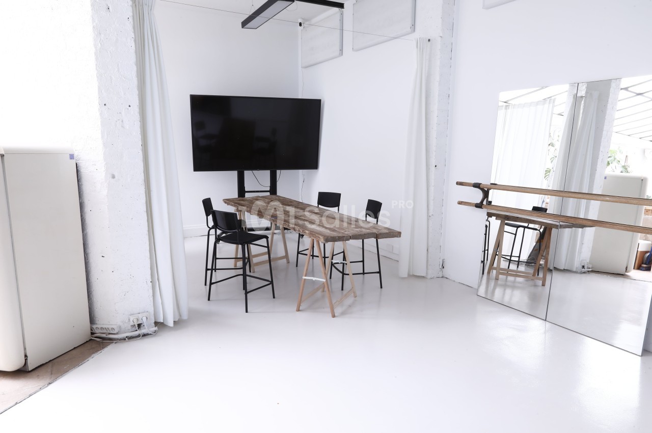 Salle lumineuse avec table en bois, chaises noires, écran mural et miroir sur le mur droit.