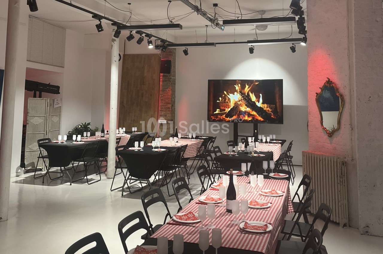 Salle aménagée pour un repas, avec tables dressées, nappes à carreaux rouges et écran affichant une cheminée.