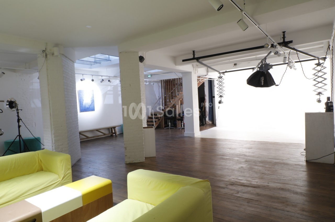 Studio photo lumineux avec fond blanc, équipements d'éclairage, parquet en bois et coin salon avec canapés jaunes.