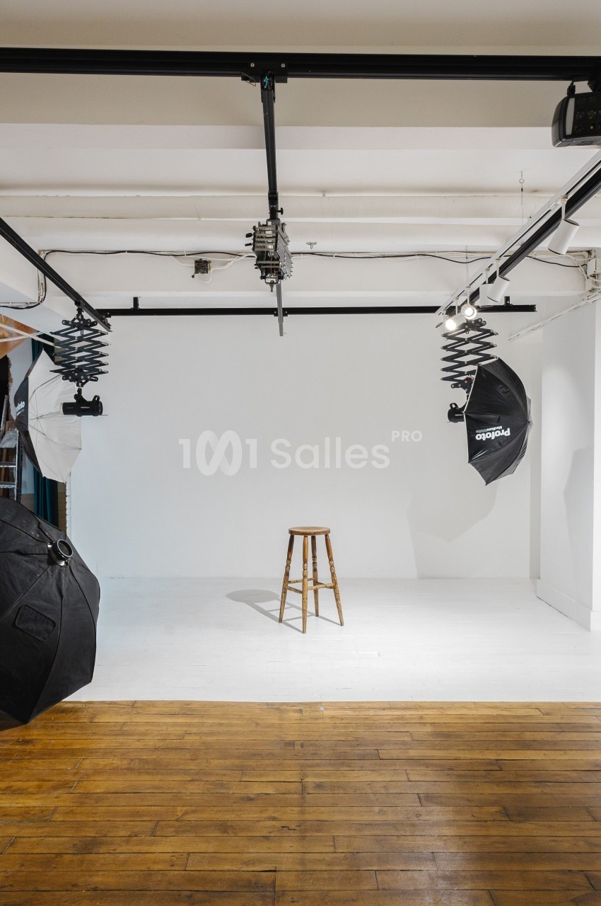Studio photo avec fond blanc, tabouret en bois au centre, éclairages et parapluies noirs suspendus.