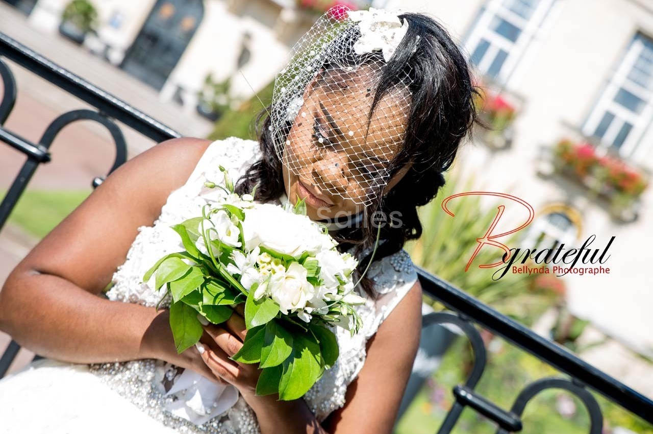 Une mariée tenant un bouquet de fleurs blanches, portant un voile en dentelle, devant un bâtiment en arrière-plan.