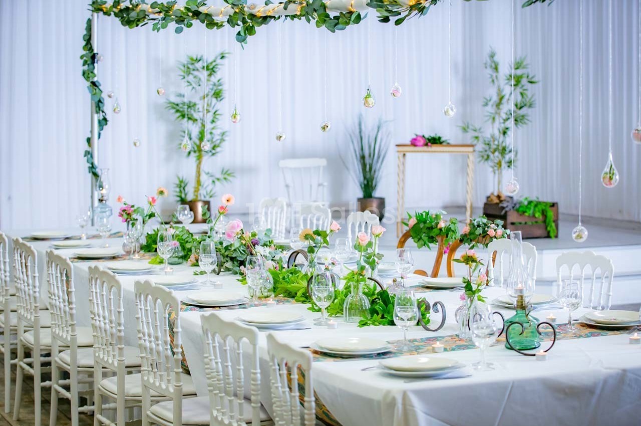 Table décorée pour un événement, ornée de plantes, fleurs et vaisselle élégante, avec fond blanc et végétation.