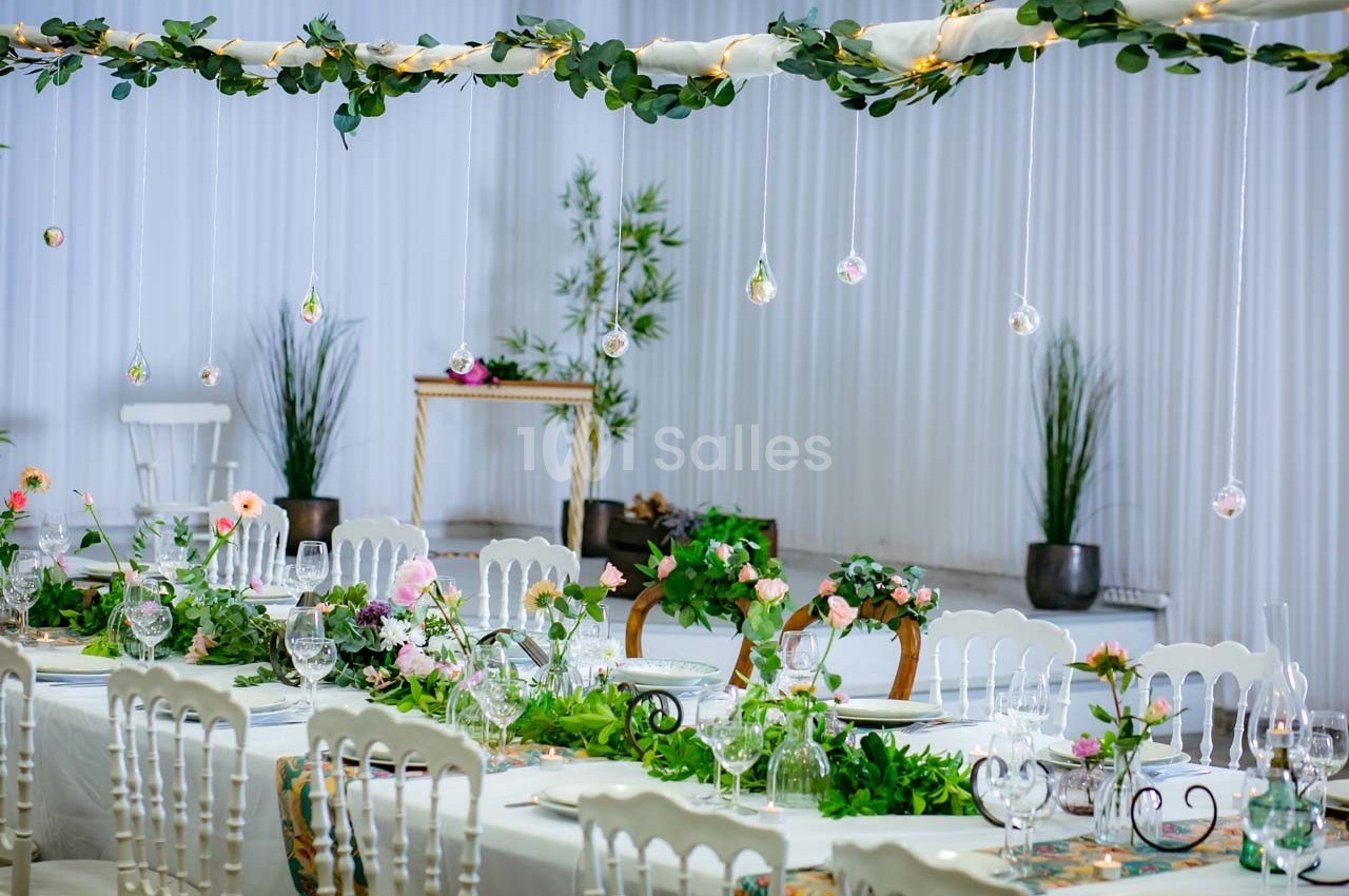 Salle de réception décorée avec des guirlandes lumineuses, des tables ornées de fleurs et une ambiance élégante.