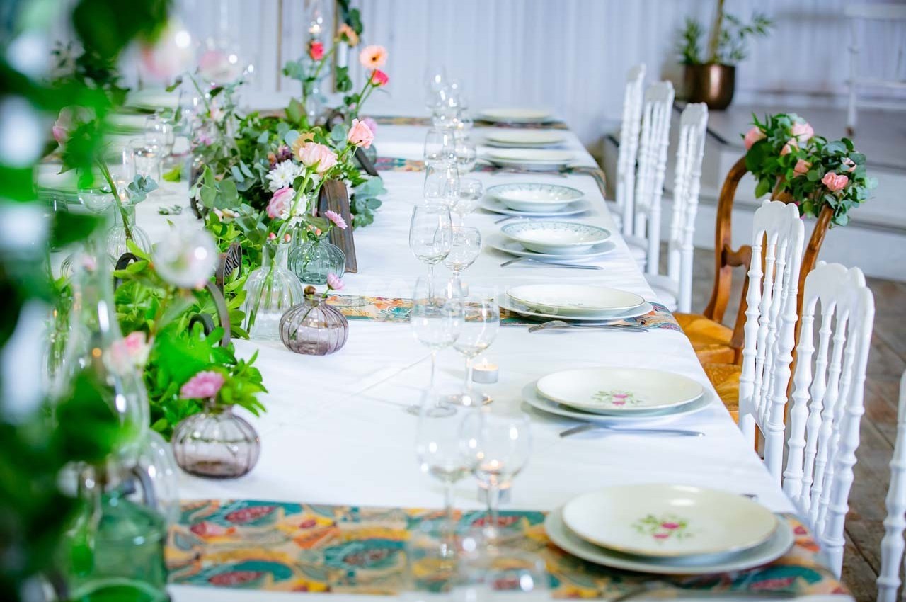 Table décorée avec des assiettes, verres et fleurs, disposée pour un repas dans un cadre élégant et lumineux.