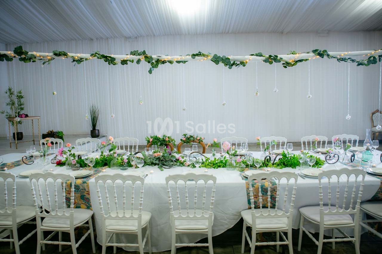 Grande table décorée de verdure, fleurs et vaisselle élégante, dans une salle lumineuse avec rideaux blancs.