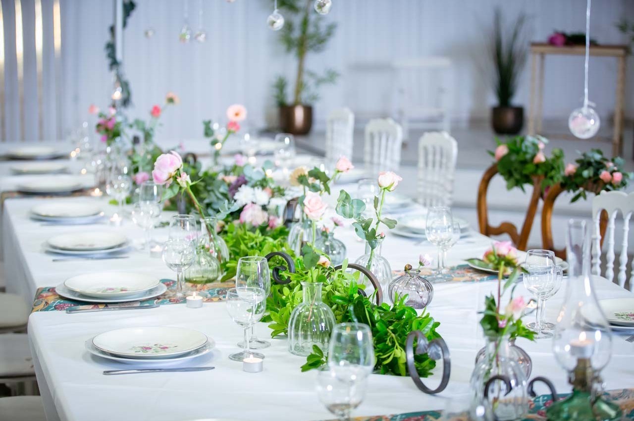 Table élégamment dressée avec nappes blanches, fleurs roses, feuillage vert et vaisselle raffinée dans une salle lumineuse.