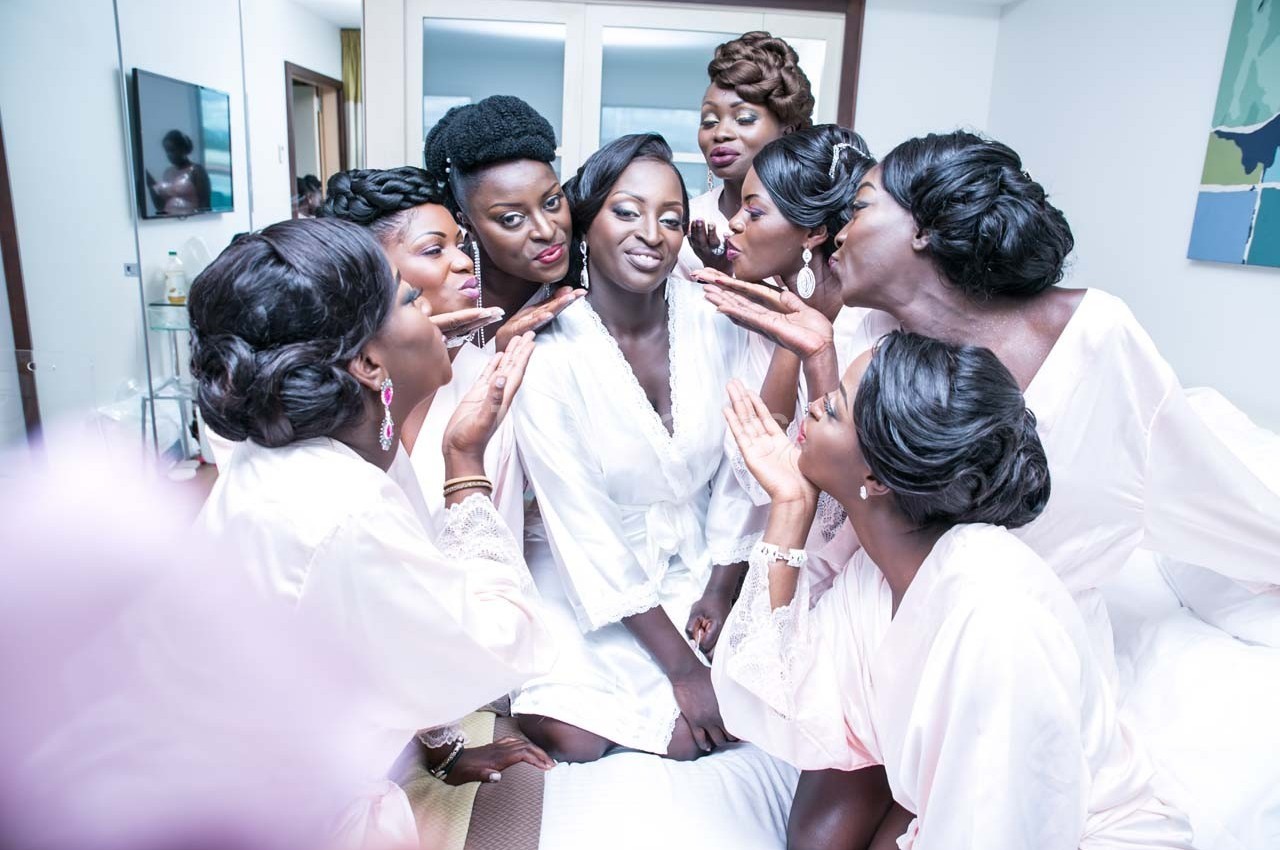 Un groupe de femmes en robes de chambre claires entoure une mariée souriante dans une chambre lumineuse.