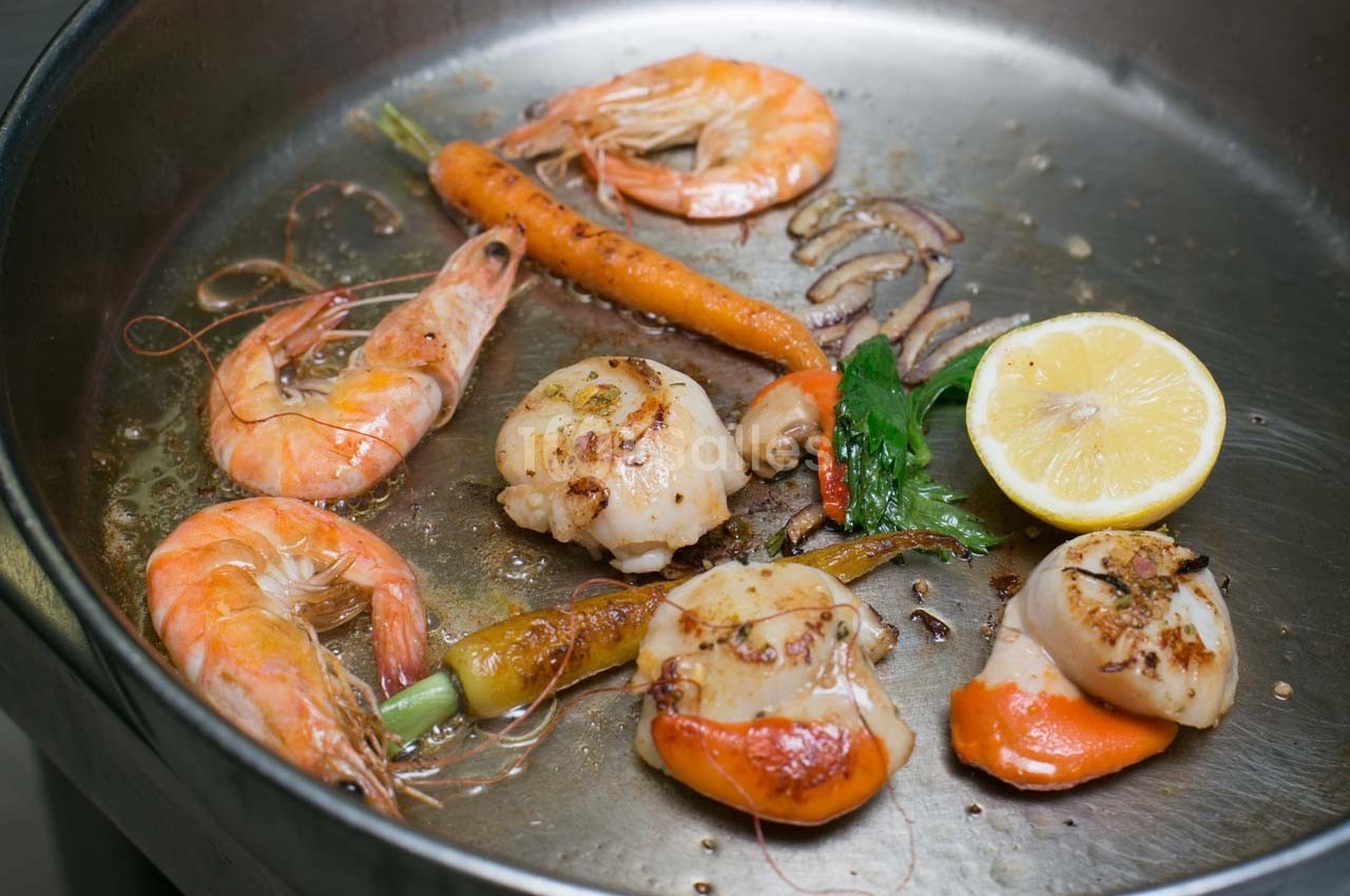 Crevettes, noix de Saint-Jacques, carottes, citron et persil cuisinés dans une poêle en métal.
