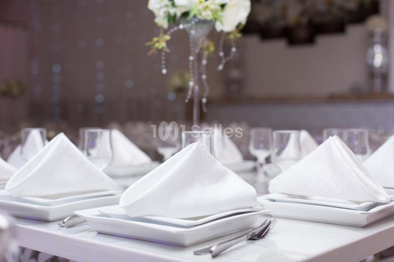 Table dressée avec des serviettes blanches pliées, assiettes empilées, couverts et verres, décor floral en arrière-plan.